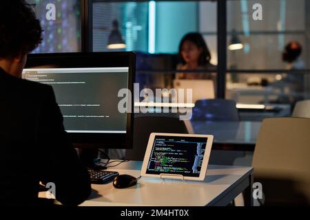 Rear view of developer sitting at table in front of computer monitor and rewrite codes from tablet pc on computer Stock Photo