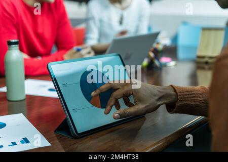 Hand of a dark-skinned man touching a digital tablet showing a pie chart on the screen. Office desk with diverse people working. Laptops, water bottle Stock Photo