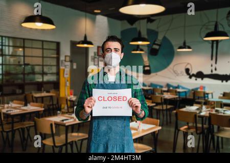 Man with a protective facial mask holds a board with sorry we are closed sign in a cafe. Concept of businesses shut down due to Covid-19 virus and qua Stock Photo