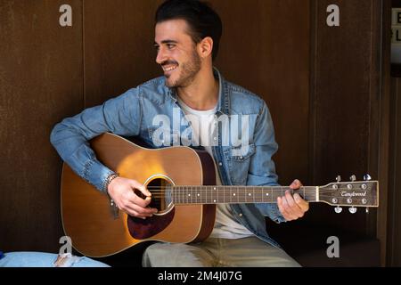 Handsome young man playing guitar. Smiling and looking away. Bearded man dressed in a denim jacket. Sitting against a brown painted wall. Enjoying, ho Stock Photo