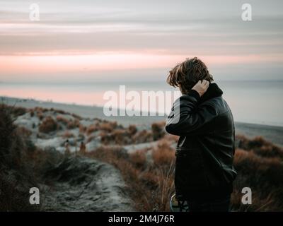 A young man wearing a black leather jacket and looking at the horizon at sunset Stock Photo