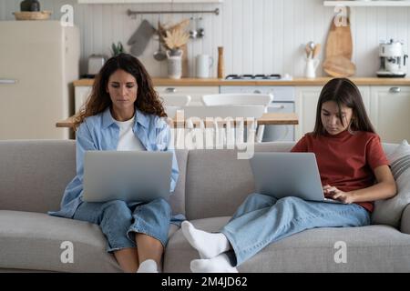 Mother and teen girl child sitting on sofa using laptops. Remote work and homeschooling Stock Photo
