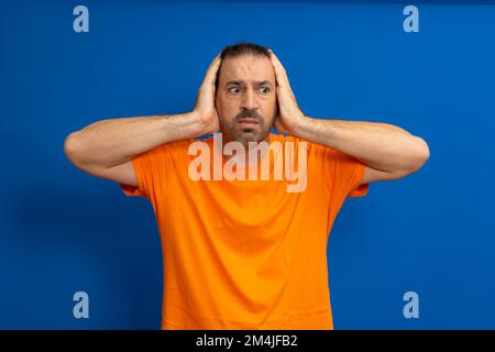 Stunned latin man wearing an orange t-shirt with his head in his hands isolated on a blue studio background. Stock Photo