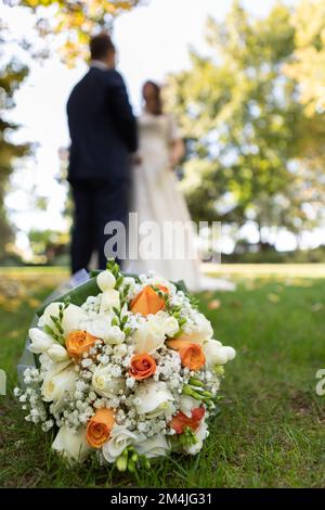 Selective focus on a bouquet of flowers in a meadow and blurry unrecognizable wedding couple in the background. Outdoor wedding in a green park Stock Photo