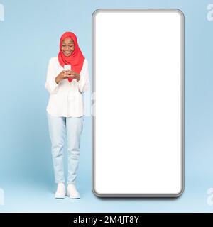 Excited black islamic woman in hijab using smartphone while standing near big cellphone with empty display, banner Stock Photo