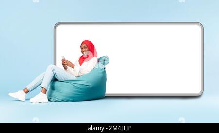 Mobile offer concept. Happy black woman in hijab sitting on beanbag chair near big smartphone with empty white display Stock Photo