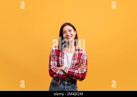 Dreamy positive asian lady standing with folded arms and looking aside, posing over yellow background, copy space Stock Photo