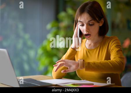 Overworked young woman checking time while having phone conversation Stock Photo