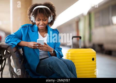 Happy Young Black Woman Messaging On Smartphone While Sitting At Railway Station Stock Photo