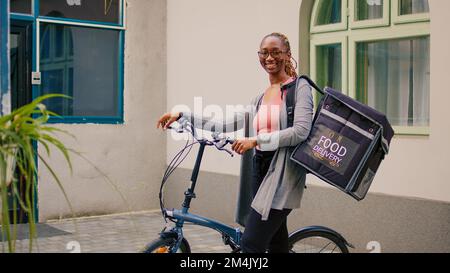 African american female carrier waiting to customer outdoors, standing next to bicycle to deliver restaurant meal order in thermal bag. Carrying backpack and holding fast food, express services. Stock Photo