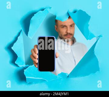 Middle aged man showing smartphone with blank empty screen through torn in blue paper wall, mockup, collage Stock Photo