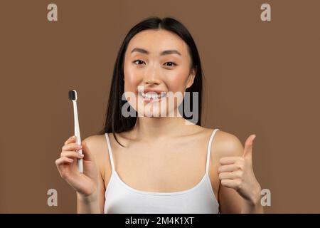 Happy asian lady holding wooden toothbrush and showing thumb up gesture, posing on brown studio background Stock Photo