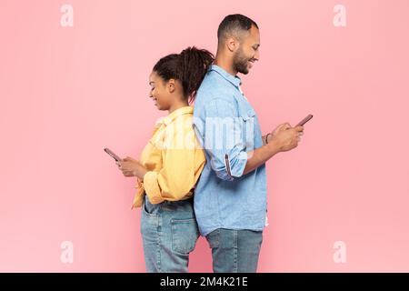 People and technology concept. Profile shot of cheerful positive black couple standing back to back, using cellphones Stock Photo