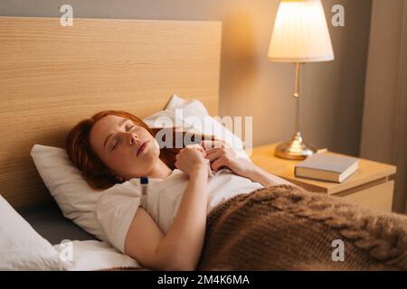 Sick tired young woman with flu lying under blanket with closed eyes on bed holding thermometer in armpit. Ill female with cold lying in bed with high Stock Photo