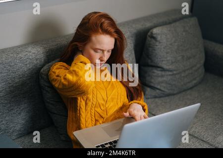 High-angle view of overworked young woman experiences severe pain in neck, rubbing it to relieve muscle tension during working on laptop computer. Stock Photo