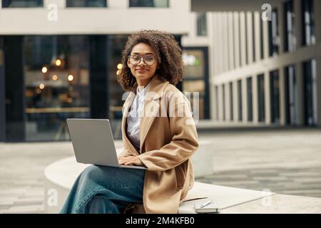 Happy african business woman working laptop while sitting outside on background of office building Stock Photo