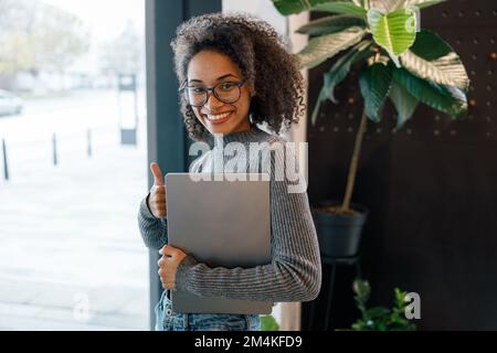 Smiling afro american woman holding laptop and looking at camera while standing near window in cafe Stock Photo