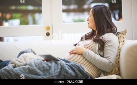 The big day is nearly here. a pregnant woman looking thoughtful while using a digital tablet at home. Stock Photo