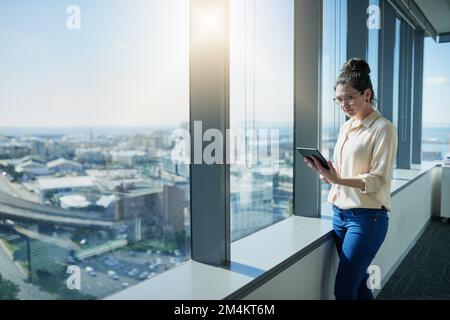 Browsing the internet from up high. a focused young businesswoman browsing on a digital tablet while standing next to a window inside of the office. Stock Photo