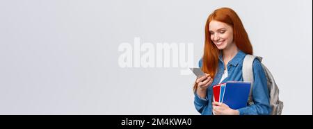 Lovely good-looking female with red hair, heading to college, holding backpack, notebooks smiling as answer friend message, chatting glancing mobile Stock Photo