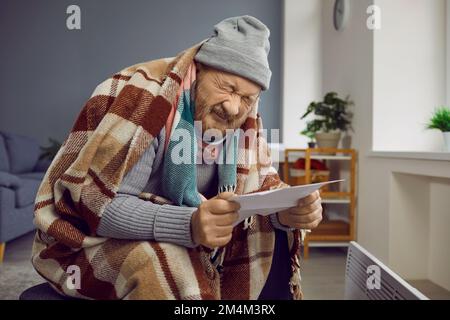 Sad poor senior man feeling cold at home, holding heating or electricity bill and crying Stock Photo