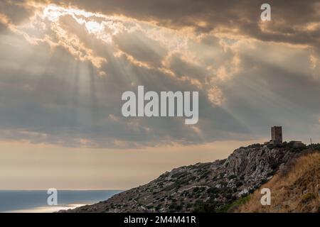 Landscape at sunset with the sun filtering through the clouds of a tower of a castle on a hill by the sea. Dark atmosphere and backlit reflections. Stock Photo