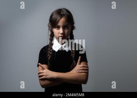 Portrait of little girl with Wednesday Addams costume during Halloween. Serious expression and dark atmosphere with dark background. Stock Photo