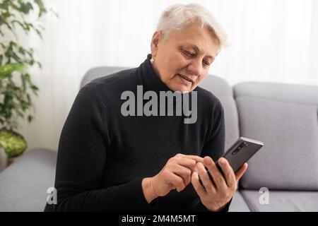 Happy mature old 60s woman holding smartphone using mobile phone app for video call, laughing while watching funny video, feeling excited winning Stock Photo