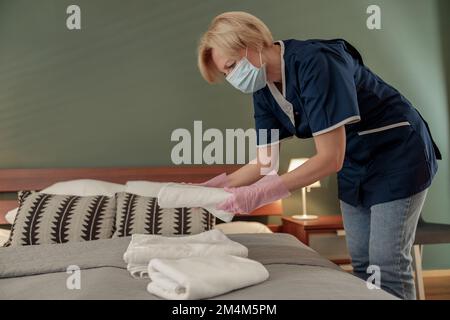 Housemaid in mask putting stack of fresh white bath towels on bed sheet. Room service concept Stock Photo