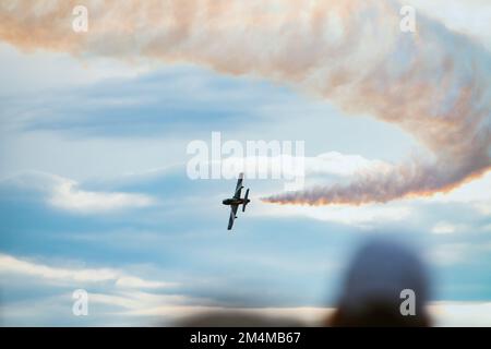 Aerial demonstration of a military airplane making a turn in the sky as it leaves a smoke trail, blurred heads of people watching Stock Photo