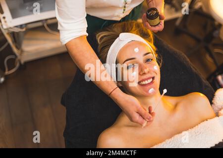 Smiling woman at beauticians. High angle portrait of young adult caucasian client looking at camera while professional SPA worker applies mask or cream on her face. High quality photo Stock Photo