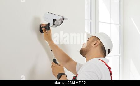 Electrician worker installs and screws video surveillance camera with backlight on wall. Stock Photo