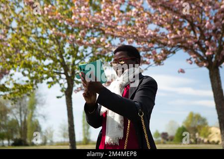 Elegant smiling African lady using smartphone in an outdoor park. Nature and flowering trees on a sunny spring day. Concept of use of technology in th Stock Photo