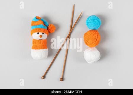 Knitted snowman in a orange blue hat and sweater with tree balls of thread and knitting needles on a gray background. Knitted Christmas decorations Stock Photo