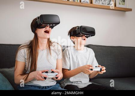Caucasian girl and her young brother having fun at home, playing driving simulation video games by using virtual reality headsets and controllers Stock Photo
