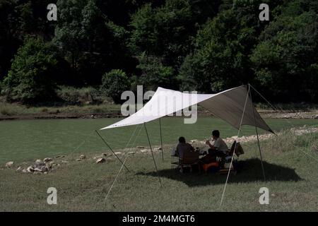 The two people sitting under the white sun shelter by the river surrounded by trees in the daytime Stock Photo