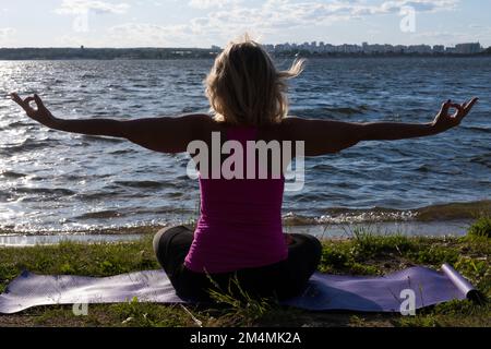 An elderly woman sitting in the lotus position on a mat in the park and looking at the lake, a view from the back. Meditation. Sports in old age. Yoga Stock Photo