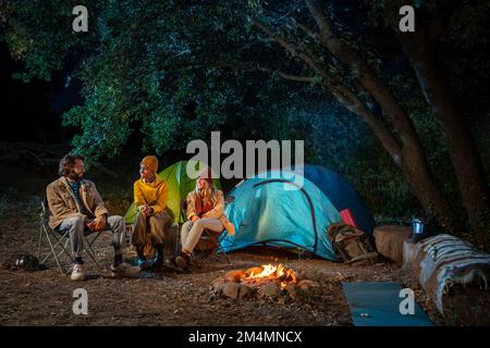 Multiethnic friends sitting around camp fire in the woods. Enjoying nature having fun together Stock Photo
