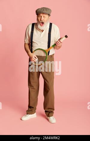Portrait of senior, old man in stylish classical clothes posing with racket, playing it like guitar over pink background. Fun Stock Photo
