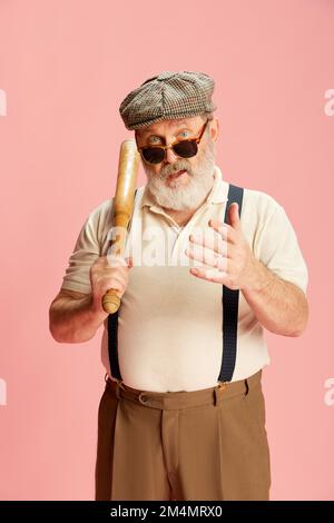 Portrait of brutal senior man in classical clothes, cap and glasses posing with bat over pink background. Questioning face Stock Photo