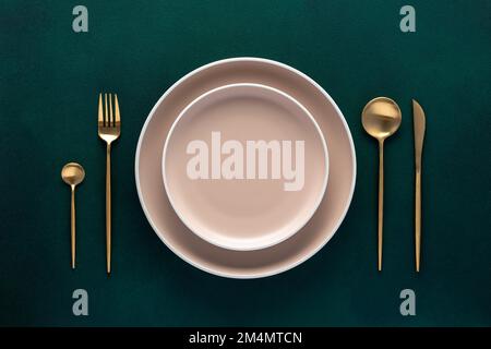 Festive place setting with beige dishes. Empty plate and gold cutlery on dark green background. Dining table in luxury restaurant. Card or menu templa Stock Photo