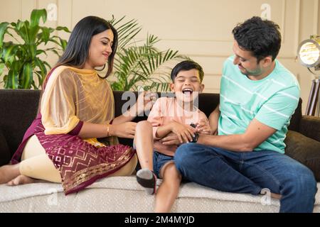 focus on mother, Happy smiling couples playing with kid by tickling or cuddling on sofa at home - concept of family bonding, affectionate and Stock Photo