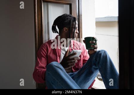 Young smiling African guy holding smartphone and cup of tea relaxing in silence by window at home Stock Photo