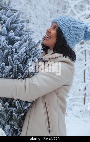 An attractive woman smiles as she holds a snowy Christmas tree in her hands as she gazes at it Stock Photo