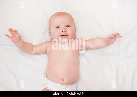 A smiling baby on a white bed. Infant baby girl spreading arms in embrace, lying on the bed on his back. Stock Photo