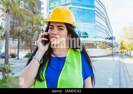 young caucasian venezuelan female engineer, wearing protective helmet and safety vest, standing outdoors talking on the phone and looking at the camer Stock Photo
