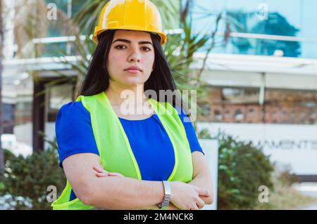 portrait of young caucasian woman of venezuelan ethnicity business engineering, wearing hardhat and safety vest, standing outdoors with arms crossed l Stock Photo