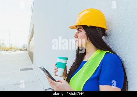 young caucasian female engineer, dressed in blue with a yellow helmet and safety vest, standing outdoors leaning against the wall with the phone in he Stock Photo