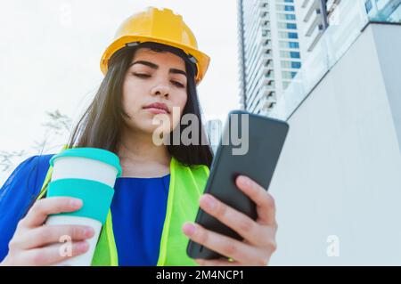 young caucasian latin woman engineer, dressed in blue with yellow hard hat and safety vest, standing outdoors drinking coffee and using phone during h Stock Photo