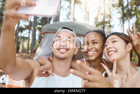 Capturing our moments outdoors. a diverse group of friends sitting together and taking selfies on a cellphone while camping in the woods. Stock Photo